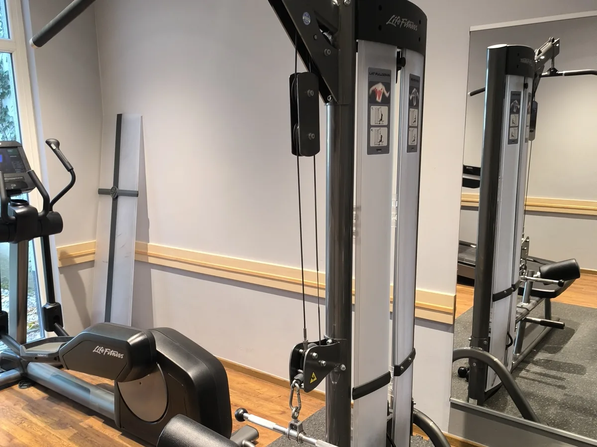 Kabelzug-Station mit offen liegenden Seilen und Umlenkrollen, daneben Crosstrainer und Rudergerät – DIN EN ISO 20957: Fitnessgeräte und stationäre Trainingsgeräte
