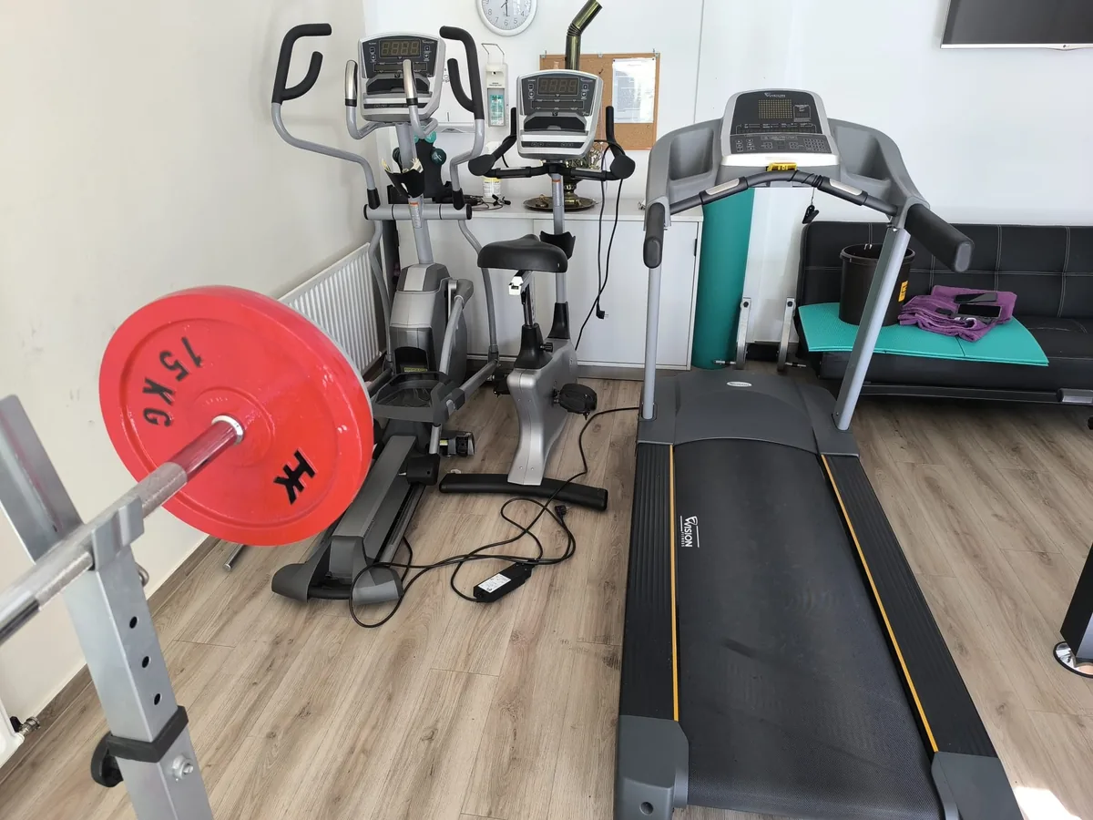 Vereinstrainingsraum mit Crosstrainer, Ergometer und Laufband, davor eine Langhantel mit Gewichtsscheibe an der Ablage – Sportgeräte in einem Vereinsheim